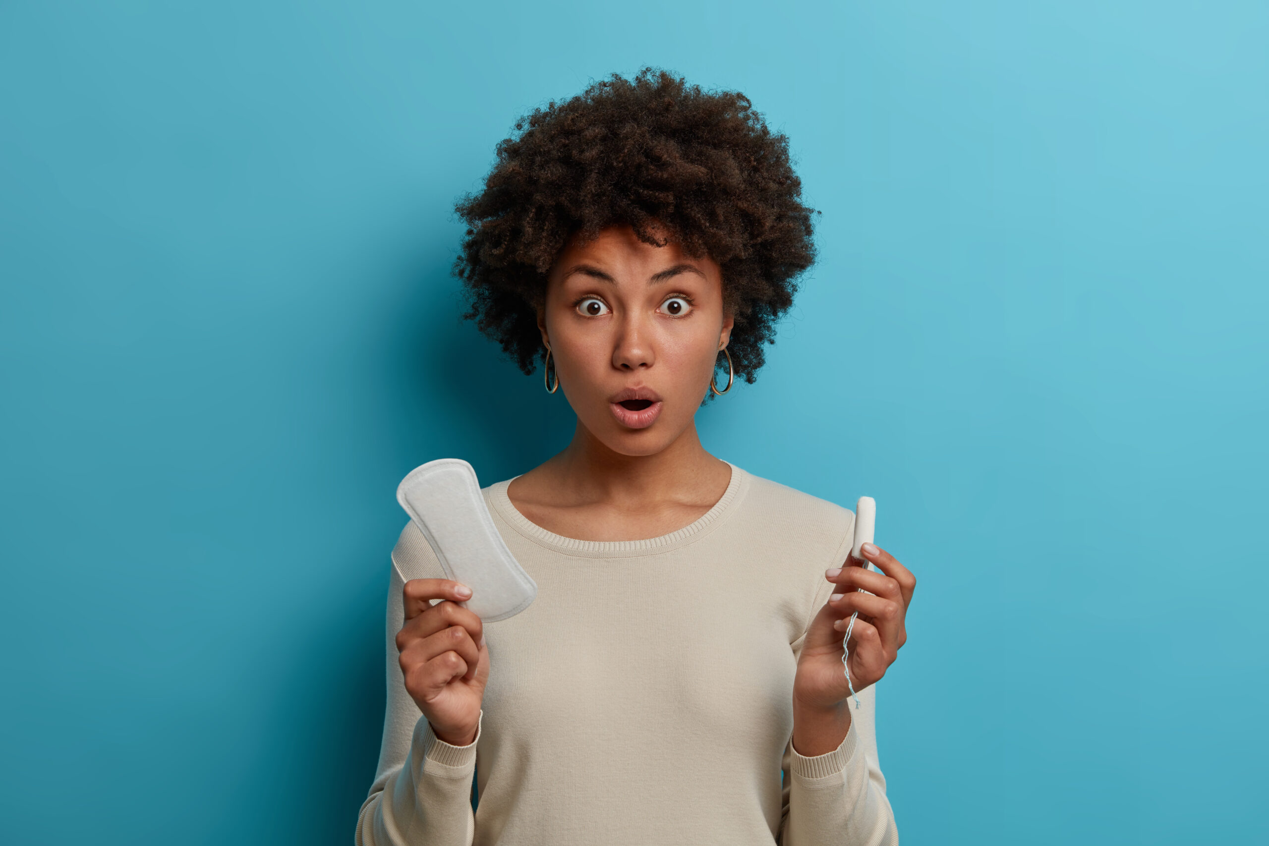 Portrait of stunned emotional young African American woman holds different types of feminine hygiene products sanitary napkin and tampon, wears casual white jumper, has periods or menstruation