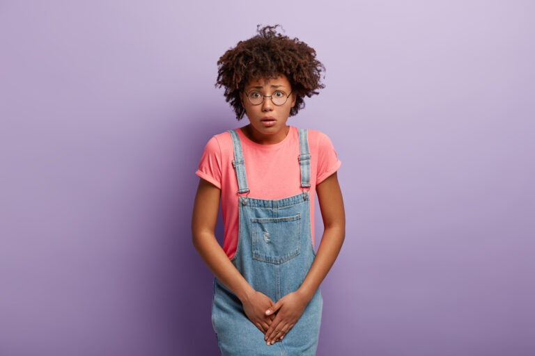 Dissatisfied girl keeps hands on crotch, presses lower abdomen, needs toilet badly, has syndrome of cystitis, wears spectacles, pink t shirt and denim sarafan, isolated on purple wall. Health problem